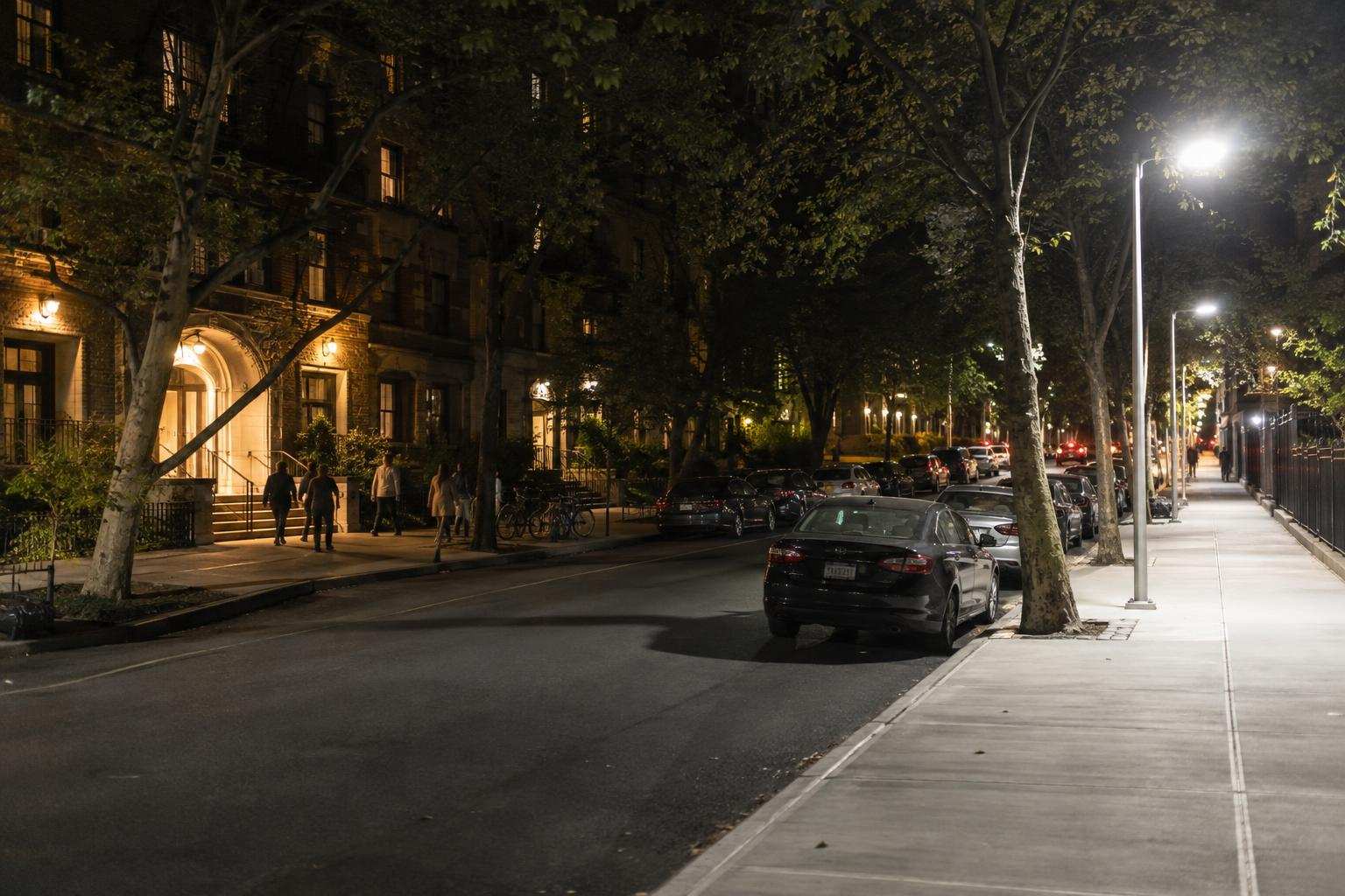 A nighttime street with a bright, nearly empty sidewalk opposite a warmer-lit sidewalk with several pedestrians.